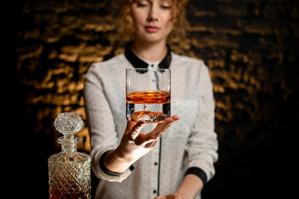 Young beautiful bartender woman hold old-fashioned glass with cold alcoholic drink. Bottle of drink stands nearby on bar counter. The kind that inspires many Drinks Buzzwords